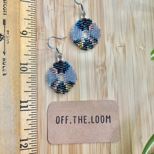 May include: A pair of silver hoop earrings with a white, blue, green and gold beaded design. The earrings are hanging from a wooden surface with a brown paper tag that says "OFF.THE.LOOM".