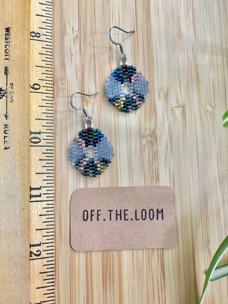 May include: A pair of silver hoop earrings with a white, blue, green and gold beaded design. The earrings are hanging from a wooden surface with a brown paper tag that says "OFF.THE.LOOM".