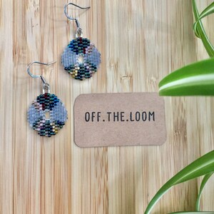 May include: A pair of silver beaded earrings with a circular design featuring a variety of colours, including blue, green, red, and gold. The earrings are on a wooden surface with a small brown card that says "OFF.THE.LOOM".