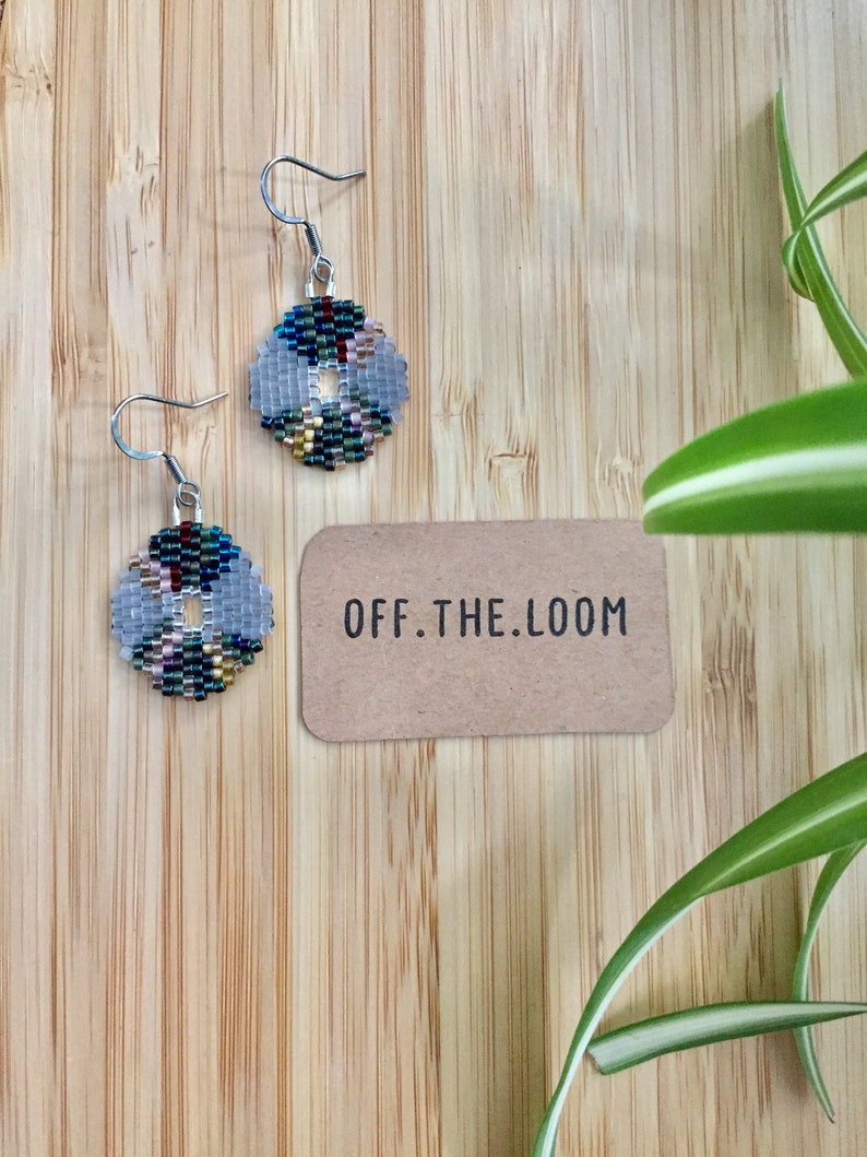 May include: A pair of silver beaded earrings with a circular design featuring a variety of colours, including blue, green, red, and gold. The earrings are on a wooden surface with a small brown card that says "OFF.THE.LOOM".