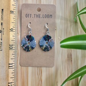 May include: A pair of silver beaded earrings with a geometric pattern in shades of green, blue, gold, and pink. The earrings are on a brown card with the text "OFF.THE.LOOM".