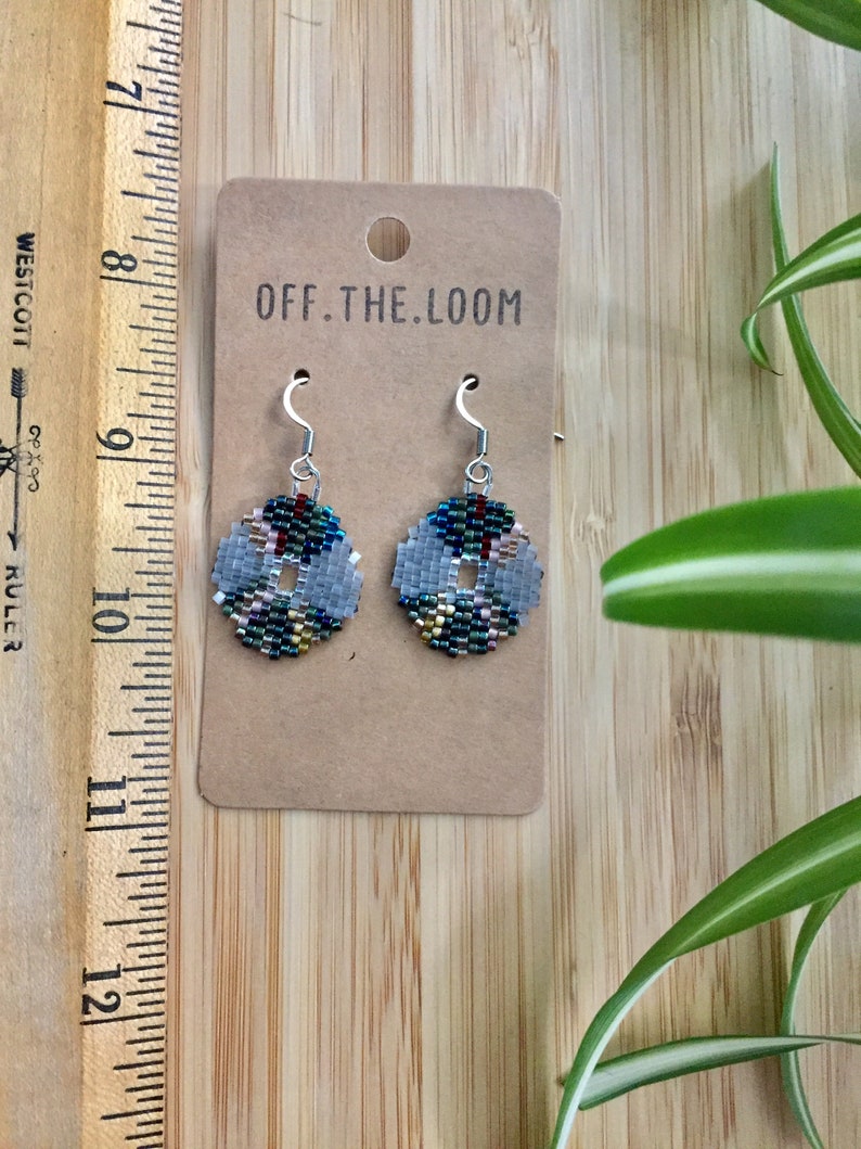 May include: A pair of silver beaded earrings with a geometric pattern in shades of green, blue, gold, and pink. The earrings are on a brown card with the text "OFF.THE.LOOM".