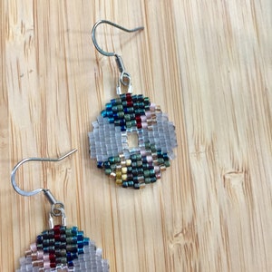 May include: A pair of silver hoop earrings with a beaded design. The beads are in shades of blue, green, gold, and red. The earrings are on a wooden surface.