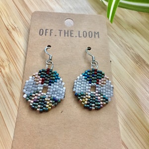 May include: A pair of beaded earrings with a white centre and a colourful, geometric pattern. The earrings are on a brown card with the text "OFF.THE.LOOM" printed on it.