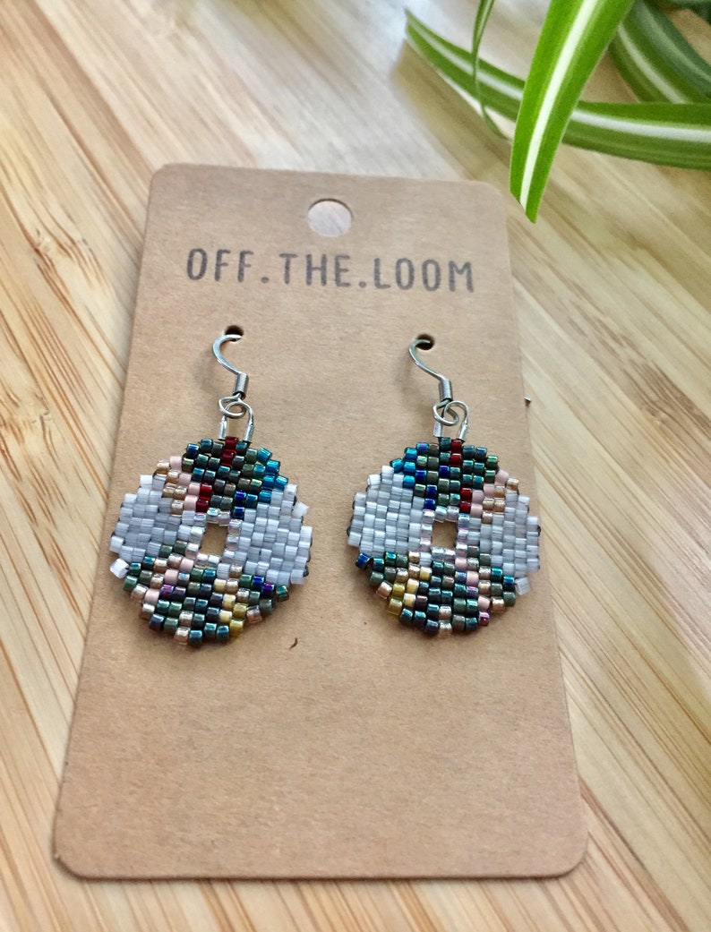 May include: A pair of beaded earrings with a white centre and a colourful, geometric pattern. The earrings are on a brown card with the text "OFF.THE.LOOM" printed on it.