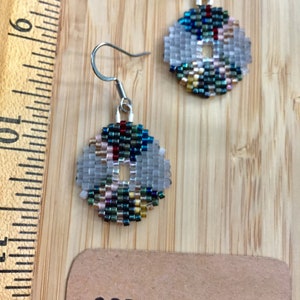 May include: A pair of silver hoop earrings with a beaded design. The beads are in shades of blue, green, gold, and white. The earrings are hanging from a wooden surface with a brown paper tag that says "OFF.THE.LOOM".