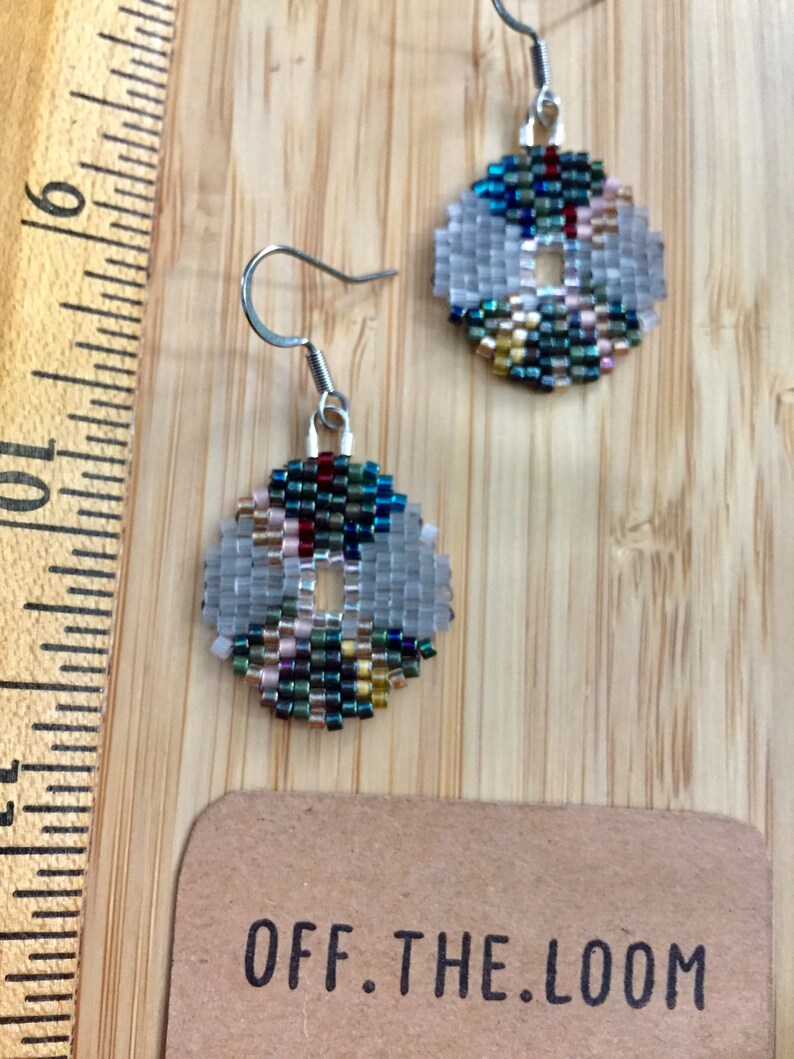 May include: A pair of silver hoop earrings with a beaded design. The beads are in shades of blue, green, gold, and white. The earrings are hanging from a wooden surface with a brown paper tag that says "OFF.THE.LOOM".