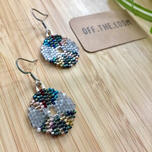 May include: A pair of silver hoop earrings with a beaded design. The beads are in shades of blue, green, gold, and white. The earrings are on a wooden surface with a brown paper tag that says "OFF.THE.LOOM".