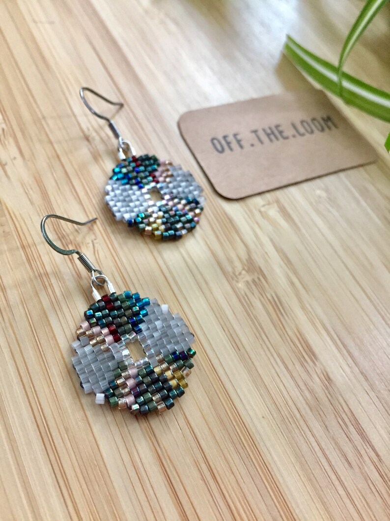 May include: A pair of silver hoop earrings with a beaded design. The beads are in shades of blue, green, gold, and white. The earrings are on a wooden surface with a brown paper tag that says "OFF.THE.LOOM".