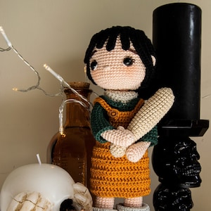 May include: A crocheted doll with black hair, a green and orange outfit, and a wooden bat. The doll is standing on a wooden surface with a white skull, cinnamon sticks, and a black candle holder with a skull design.