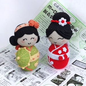 May include: Two crocheted dolls dressed in traditional Japanese clothing. The doll on the left is wearing a green and yellow kimono with a white sash. The doll on the right is wearing a red and white kimono with a white sash. Both dolls have black hair and smiling faces.