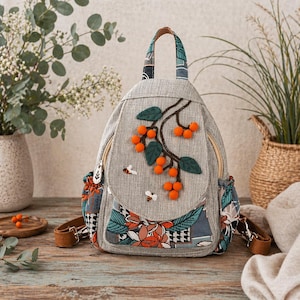 May include: A gray backpack with a flap featuring an embroidered branch with orange berries and green leaves. The backpack has a patterned handle and side pockets, with two embroidered bees. The straps are brown.