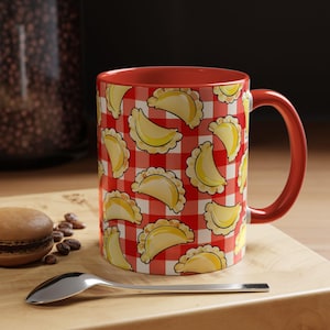 May include: A ceramic mug with a red and white checkered pattern featuring a repeating illustration of yellow pierogi. The mug has a solid red interior and a matching red handle, providing a vibrant contrast to the patterned exterior surface.