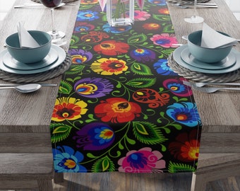 May include: A black table runner with a colorful floral pattern. The pattern features red, yellow, blue, green, and purple flowers with intricate details. The runner is laid out on a wooden table with place settings.