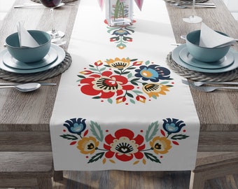 May include: White table runner with a floral pattern in red, blue, yellow, and green. The pattern features stylized flowers and leaves. The runner is on a wooden table with two place settings.