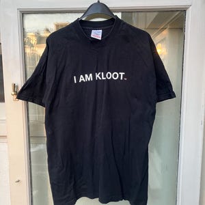 May include: Black t-shirt with the text "I AM KLOOT" in white, centered on the chest. The shirt is hanging from a black hanger against a white door and window background. The shirt appears to be made of cotton.