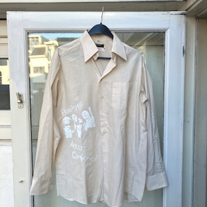 Puede incluir: Una camisa beige de manga larga con botones y un estampado gráfico blanco de una banda llamada "Bloodshot" y el texto "Adult Commitment".