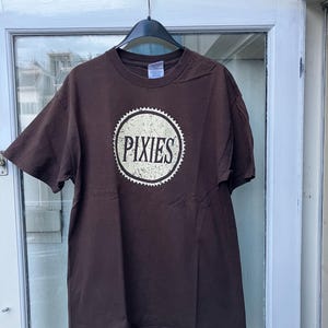 May include: A brown t-shirt with a white circle logo that says "PIXIES" in a vintage style font.
