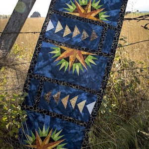 May include: A blue quilted table runner with a geometric design. The runner features repeating starburst and triangle patterns in shades of orange, green, and white. The background is a mottled blue fabric with a black patterned border. The runner is draped over a wooden fence.