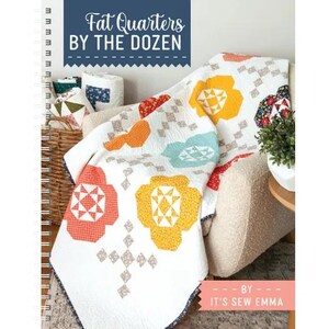 以下が含まれることがあります： 「Fat Quarters by the Dozen」というタイトルのスパイラル綴じの本。オレンジ、黄色、青色の大きな花柄が特徴の白いキルト。キルトはクリーム色の肘掛け椅子に掛けられています。追加の生地ロールとバスケットが見えます。