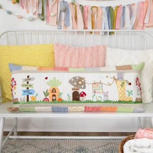 May include: A long, rectangular pillow with an embroidered design of a bunny rabbit village. The pillow features a carrot house, a mushroom house, a sign that says "Hoppin' Down the Bunny Trail", and a bunny rabbit in a yellow rain boot. The pillow is on a white bench with a yellow, pink, and white throw pillows behind it.