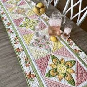 May include: A quilted table runner with a floral and geometric pattern in shades of pink, green, yellow, and white. The runner is placed on a wooden table, with a glass pitcher, glasses, and lemons visible.