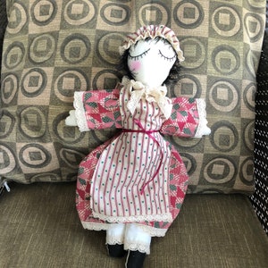 Puede incluir: Una muñeca de trapo hecha a mano con cabello negro, ojos cerrados y un vestido rosa y blanco. La muñeca está acostada sobre una almohada con estampado marrón y blanco.