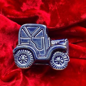 Puede incluir: Pequeña figura de coche antiguo de cerámica azul, con ruedas detalladas y diseño de techo. El coche se encuentra sobre un fondo de terciopelo rojo vibrante, creando un contraste llamativo. Una pieza coleccionable encantadora.