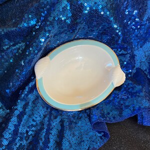 May include: A white ceramic dish with a light blue rim and gold trim. The dish has two shell-shaped handles on the sides. The dish is sitting on a blue sequined fabric.