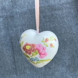 Może przedstawiać: Biała ceramiczna ozdoba w kształcie serca z kwiatowym wzorem przedstawiającym różowe, czerwone i niebieskie kwiaty oraz zielone łodygi. Ozdoba wisi na jasnoróżowej wstążce przymocowanej do zakrzywionego metalowego haczyka w kolorze brązu.