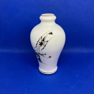 May include: A small, white ceramic vase with a narrow neck and a rounded body. The vase features a hand-painted design of black and brown grass and flowers with red accents. Gold trim accents the top and bottom.