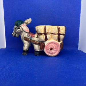 Puede incluir: Una figura de cerámica de un burro tirando de un carro con un recipiente hueco. El burro es gris con una gorra verde y orejas rosadas. El carro es de color crema con líneas de cuadrícula marrones y una rueda rosa con diseño floral.