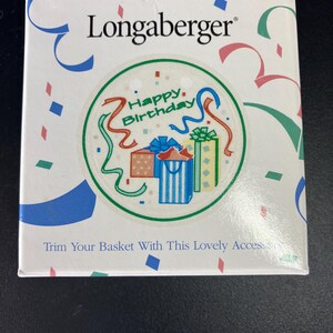 Puede incluir: Una caja blanca de Longaberger con un diseño circular con las palabras "Happy Birthday" e ilustraciones de regalos envueltos. La caja está decorada con confeti y cintas azules, verdes y rojas.