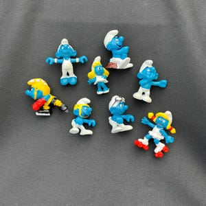 Puede incluir: Colección de figuras de los Pitufos en varias poses. Las figuras son principalmente azules con sombreros blancos y detalles de ropa. Algunos Pitufos se representan en poses de acción, mientras que otros están de pie o sentados.