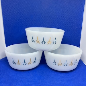May include: Three white, vintage Pyrex bowls. Each bowl has a rounded shape and is decorated with a repeating pattern of stylized starbursts in teal and gold. The bowls are arranged on a blue surface.
