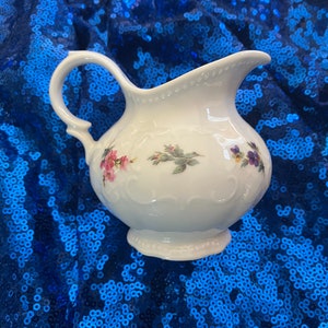 Può includere: Un creamer in porcellana bianca con un motivo floreale. Il creamer ha un manico e un beccuccio. È decorato con fiori rosa, viola e verdi.