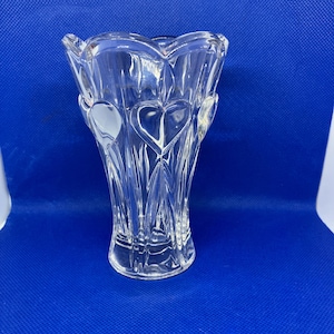 May include: Clear glass vase with a heart-shaped design. The vase has a flared top and a textured, ridged body. The heart shapes are visible throughout the vase, adding a decorative element. The vase is on a blue background.