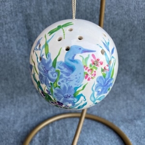 May include: A spherical white ceramic ornament featuring a hand-painted blue bird surrounded by blue flowers, green foliage, and a small green dragonfly. The top of the sphere has several small circular perforations and a thin gold hanging cord.