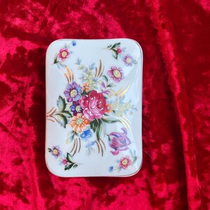 May include: A rectangular white porcelain trinket box featuring a central floral arrangement with pink, purple, orange, and blue flowers, accented by green leaves and delicate gold painted flourishes across the surface of the lid.