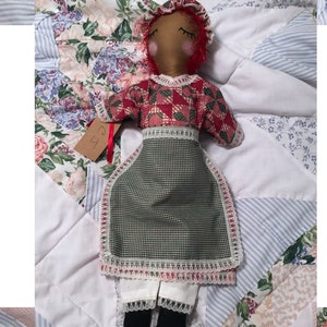 Puede incluir: Una muñeca de trapo hecha a mano con un vestido a cuadros rojo y verde y un delantal blanco. La muñeca tiene un gorro de pelo rojo y los ojos cerrados. La muñeca está tumbada sobre una colcha con estampado floral.
