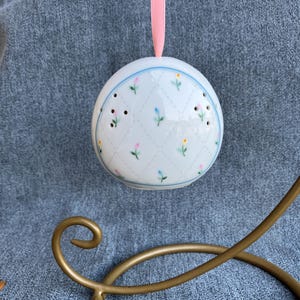 May include: A white ceramic egg-shaped ornament featuring a delicate diamond grid pattern with small hand-painted pink, yellow, and blue flowers. The ornament has small ventilation holes and is suspended by a light pink ribbon from a gold metal stand.