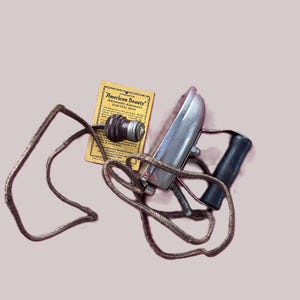 Puede incluir: Una plancha eléctrica antigua con base plateada y mango negro. Una tarjeta amarilla con el texto "American Beauty" está junto a la plancha. La plancha tiene un cable marrón.