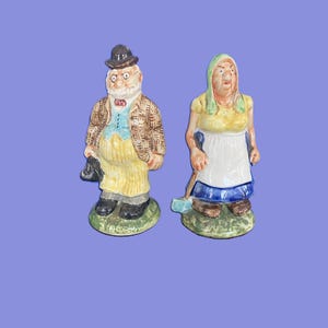 May include: Two ceramic figurines of an elderly couple. The man wears a black hat, glasses, and a tweed jacket. The woman wears a headscarf and apron, holding a broom. Both stand on green bases.