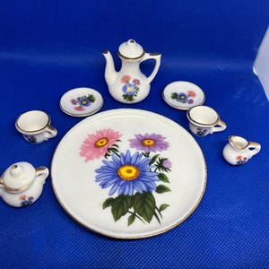 Puede incluir: Un juego de té de porcelana en miniatura con un diseño floral. El juego incluye una tetera, una jarra de crema, un azucarero, dos tazas, dos platillos y un plato grande. Las flores son rosas, azules y moradas con ribetes dorados.