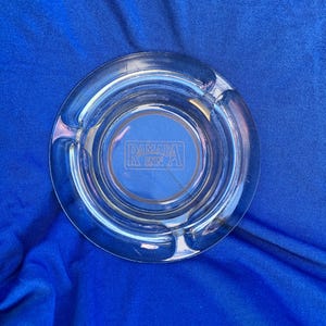 May include: A clear glass ashtray with a circular design. The ashtray has a recessed centre with the text "RAMADA INN" in a rectangular frame. It features three notches for cigarettes. The ashtray is on a blue surface.