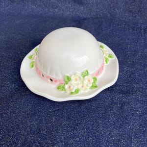 May include: A white ceramic hat-shaped trinket dish featuring a pink ribbon band with small cutouts. The brim is decorated with a cluster of sculpted white flowers with yellow centres and green leaves, creating a detailed floral accent on the side.