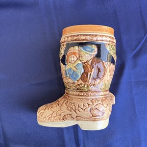 May include: A ceramic boot-shaped mug with a brown, tan, and blue glaze. The mug features a raised design of two people in traditional clothing.