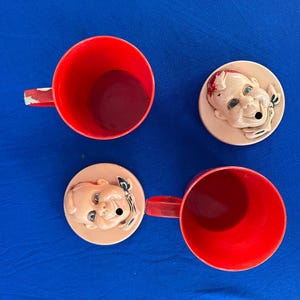 May include: Two red plastic mugs with handles and two matching lids. Each lid features a smiling baby face design. The mugs and lids are arranged on a blue surface, creating a playful and vintage aesthetic.