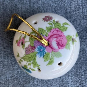 Puede incluir: Una bola de cerámica blanca redonda con un patrón floral de rosas rosas, flores azules y hojas verdes. El objeto tiene varios orificios de ventilación circulares pequeños e incluye un cordón dorado para colgarlo.