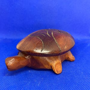 Puede incluir: Una figura de tortuga de madera tallada con un caparazón pulido de color marrón oscuro que presenta detalles de líneas grabadas. La tortuga tiene un cuerpo liso y redondeado, una cabeza prominente y cuatro patas cortas de madera.